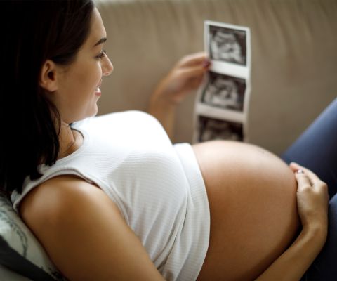 Schwangere schaut lächelnd auf Ultraschallbilder, mit einer Hand auf dem Bauch
