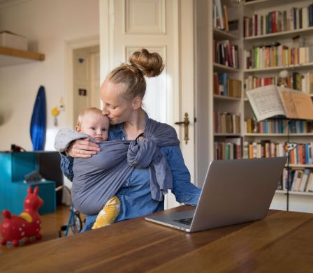 Maman portant son bébé dans une écharpe porte-bébé tandis qu’elle travaille sur l’ordinateur portable
