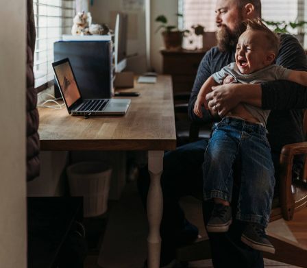 Ein Vater hat sein weinendes Kind im Homeoffice auf dem Schoss und ist am Laptop